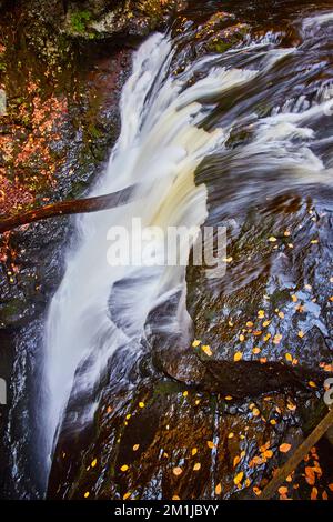 In der Nähe des Wasserfalls, der in die Tiefe ragt, mit Herbstblättern, die auf Felsen ruhen Stockfoto