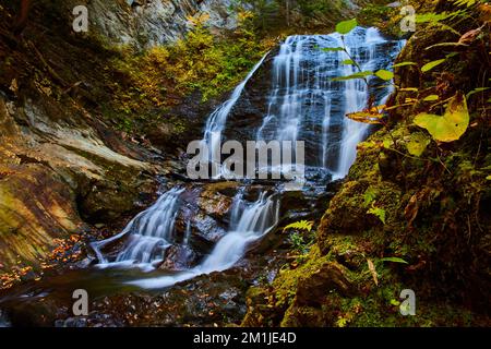 Großer tosender Wasserfall in einer engen Schlucht, umgeben von moosbedeckten Felsen und herabfallenden Blättern Stockfoto