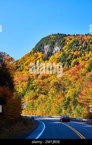 Wunderschöne Laubbäume bedecken felsige Berge, die von der Straße aus betrachtet werden, während ein Auto vorbeifährt Stockfoto