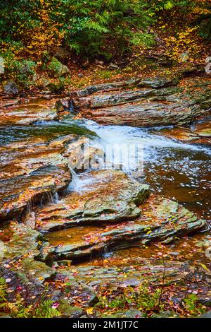 Kleiner Wasserfall, der sich durch den Fluss stürzt, mit herbstlichen Blättern, die die herbstlichen Felsen bedecken Stockfoto