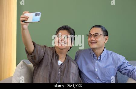 Ein asiatisches Paar verbringt Zeit miteinander im Wohnzimmer. Frau und Mann im Ruhestand machen Selfie-Fotos mit dem Handy. Moderne Lebensweise Menschen Beziehung A Stockfoto