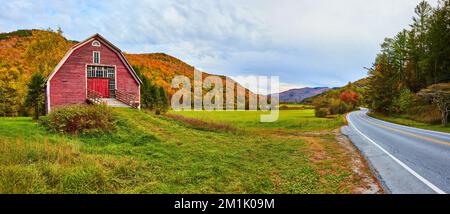 Panoramablick von der Straße in Vermont auf die rote Scheune auf grasbedeckten Feldern, umgeben von Herbstbergen Stockfoto