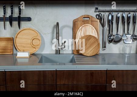 Arbeitsbereich der Küche. Küchenarbeitsplatte mit eingebautem Spülbecken, Messer an der Wand und Schneidebretter stehen darauf in einer dunklen Küche. Stockfoto