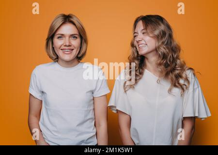 Positiv zwei junge Frauen haben Spaß, eng nebeneinander zu stehen, gekleidet, zwanglos isoliert und mit orangefarbenem Hintergrund. Lebensstil der Menschen. Stockfoto