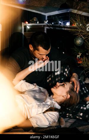 Frohe Weihnachten. Ein junges Paar, das Weihnachten oder Silvester zu Hause feiert. Ein romantisches Paar im Pyjama, das sich zu Weihnachten umarmt Stockfoto