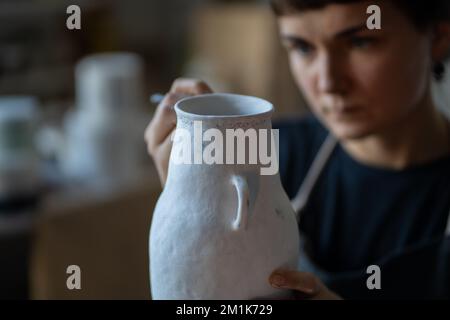 Fokussierte Brünette Frau malt Text mit Pinsel auf weiße Keramikvase am Tisch auf verschwommenem Hintergrund Stockfoto