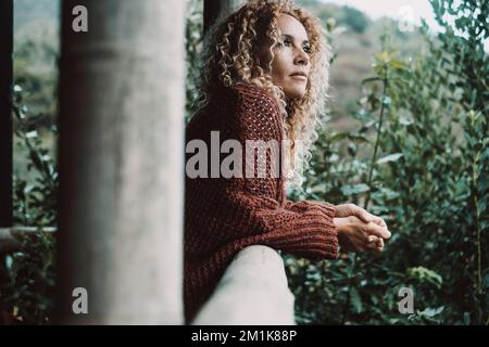 Seitlicher Porträtblick einer attraktiven jungen, reifen Frau, die sich auf der Terrasse mit grünem Naturgarten und Wald im Hintergrund im Freien aufhält. Konz Stockfoto