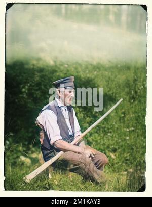 Vintage-Farbfoto eines alten Mannes, der an einem sonnigen Sommertag auf einem Feld saß und eine alte Militärmütze trug, die ein Ruder hielt. Ungefähr 1900 Stockfoto