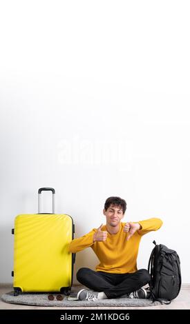 Junger Reisender, der Daumen hoch und Daumen runter macht, unterschreibt mit dem Koffer auf isoliertem weißem Hintergrund. Vertikales Banner Stockfoto
