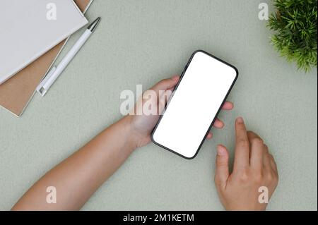Eine weibliche Hand, die ein Smartphone-Modell mit weißem Bildschirm über ihren modernen, minimalistischen Büroschreibtisch hält. Graue Ansicht des Arbeitsplatzes von oben mit weiblichen Händen, die das Telefon halten. Stockfoto