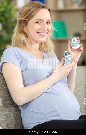Schwangere Frau zeigt Neugeborenenschuhe im Wohnzimmer Stockfoto