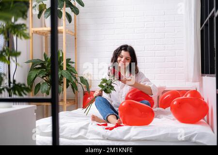 Mädchen in der Liebe mit Blumenstrauß auf dem Bett rote Herz Form Ballons. Die Frau freut sich über das Geschenk am Valentinstag. Easy-going Mädchen mit langen Haaren Stockfoto