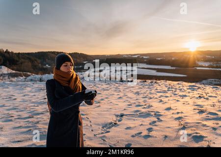 Eine junge Frau mit Mantel und Hut steht bei Sonnenuntergang auf einem schneebedeckten Feld und hält eine Handvoll Schnee in ihren Händen Stockfoto