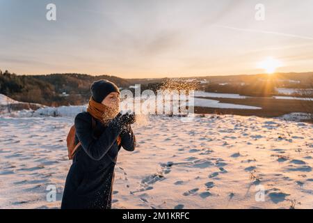 Eine Frau bläst im Winter Schnee von ihren Handflächen auf einem schneebedeckten Feld Stockfoto