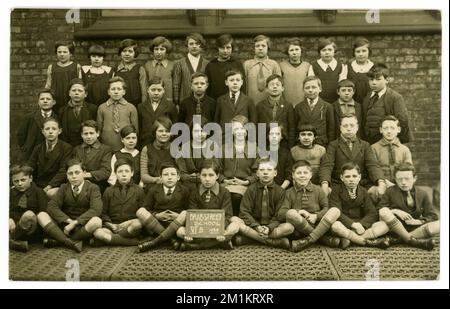 Originalpostkarte der Brae Street School aus den 1920er Jahren aus dem Jahr V1B, ausgestellt von 1928 (auf dem Schild). Portrait der Gruppenklasse draußen auf dem Schulspielplatz, in gemischten Schuluniformen und anderen Moden. Auf der Rückseite ist das Alter von 12 Jahren geschrieben. Liverpool, Merseyside, England, Großbritannien Stockfoto