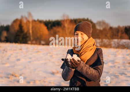 Eine lächelnde Frau mit Schal und Hut hält im Winter eine Handvoll Schnee Stockfoto