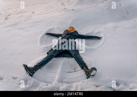 Eine junge Frau macht einen Schneeengel, der im Schnee liegt und Arme und Beine seitlich gespreizt hat Stockfoto