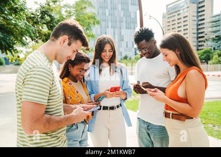 Multirassische Gruppe junger, lächelnder Schüler, die sich mit ihren Handys amüsieren. Freunde im Teenageralter, die sich nach dem Unterricht auf dem Campus SMS schicken. Hochwertiges Foto Stockfoto