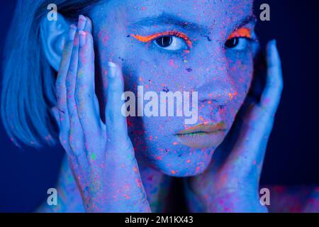 Porträt der Frau in Neon-Make-up und fluoreszierende Farbe, die Hände in der Nähe des Gesichts hält auf dunkelblau, Stock-Bild Stockfoto