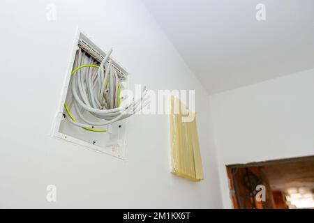 Ein Bündel verschiedener Kabel hängt an einem offenen Sicherungskasten aus Kunststoff, der an der Putzwand montiert ist, und der Stromzähler ist mit Klebeband abgedeckt Stockfoto