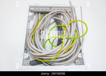 Ein Bündel verschiedener Kabel hängt an einem Kunststoff-Sicherungskasten, der an der Putzwand montiert ist, Gebäude im Bau, laufende Arbeiten. Installation von Elektro Stockfoto