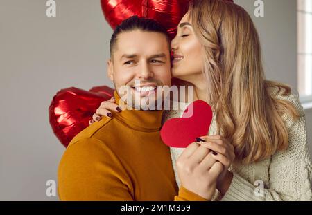 Glückliche Frau umarmt ihren Mann, küsst ihn und gibt ihm eine rote herzförmige Valentinskarte Stockfoto