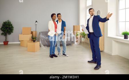 Professioneller männlicher Immobilienmakler zeigt jungen afroamerikanischen Paar eine helle neue Wohnung. Stockfoto