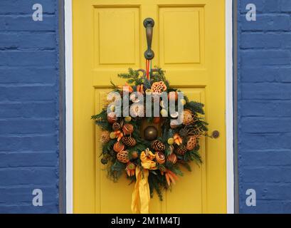 Ein wunderschöner Weihnachtskranz mit Tannenzapfen und Orangen hängt an einer gelben Tür, umgeben von blauen Ziegeln Stockfoto