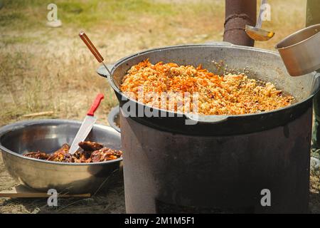 An einem sonnigen Tag im Freien wird traditioneller orientalischer Pilaf in einem großen Kessel gekocht. Echter usbekischer Pilaf in der Steppe Stockfoto