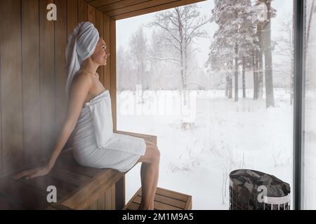 Eine Frau mit weißem Handtuch, die sich in der Sauna entspannt und die Winterlandschaft genießt Stockfoto