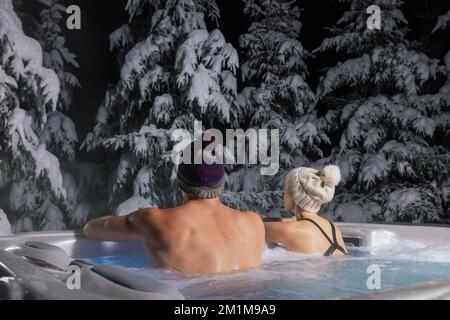 Ein Paar, das sich im Wannen-Whirlpool im Freien entspannen und die verschneite Winterwaldlandschaft im Spa Resort genießen kann Stockfoto