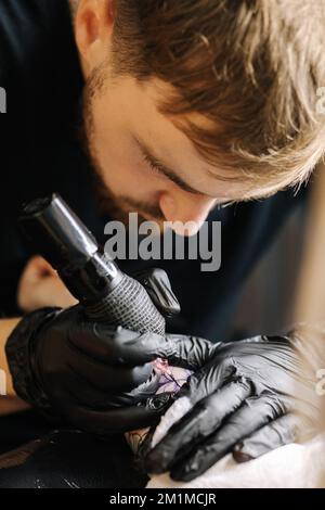 Bei der Nahaufnahme eines jungen professionellen Tätowierers wird schwarze Tinte mit einer Nadel aus einem kabellosen Tätowiergerät in die Haut eingeführt. Tattoo auf der Hand. Stockfoto