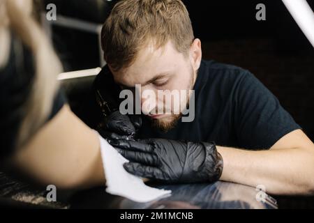 Bei der Nahaufnahme eines jungen professionellen Tätowierers wird schwarze Tinte mit einer Nadel aus einem kabellosen Tätowiergerät in die Haut eingeführt. Tattoo auf der Hand. Stockfoto