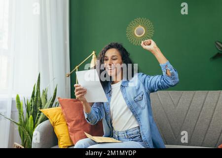 Die glückliche Frau zu Hause erhielt einen netten Benachrichtigungsbrief und ein glückliches Lächeln, eine hispanische Frau zu Hause mit einem Briefumschlag, der auf dem Sofa im Wohnzimmer saß. Stockfoto
