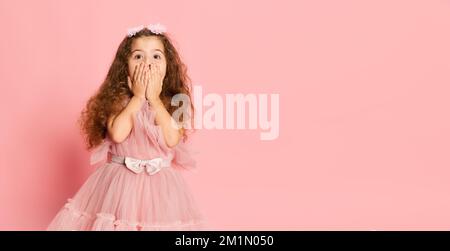 Das Ist Toll. Ein emotional süßes kleines Mädchen, ein Kind mit langen Locken und festlichem Prinzessinnenkleid, isoliert auf pinkfarbenem Hintergrund. Glückliche Kindheit, Kind Stockfoto