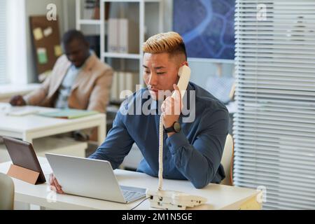 Porträt eines asiatischen Mannes, der im Büro über ein schnurgebundenes Telefon telefoniert und einen Laptop benutzt Stockfoto