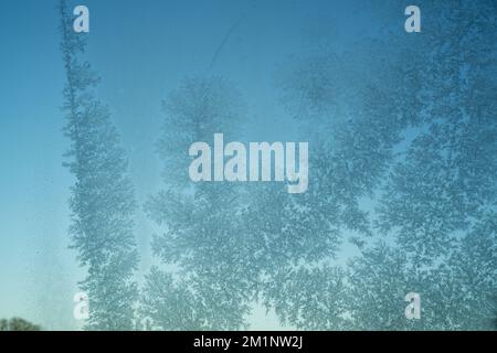 Frostmuster auf der Glasscheibe. Milchglas mit blauem Himmel dahinter. Empfindliche Kristalle, die sich bei kaltem Wetter bilden. Stockfoto