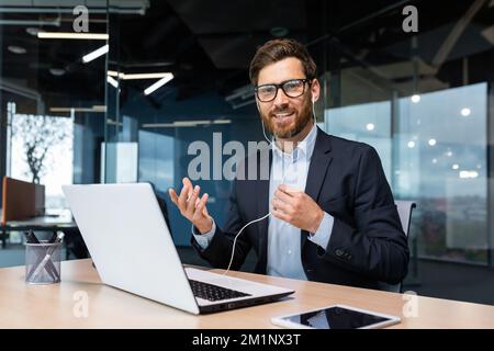 Ein junger Geschäftsmann sitzt mit Kopfhörern im Büro, leitet ein Geschäftstreffen, eine Konferenz über ein Videogespräch mit einem Laptop. Er winkt mit den Händen, sieht in die Kamera, lächelt. Stockfoto