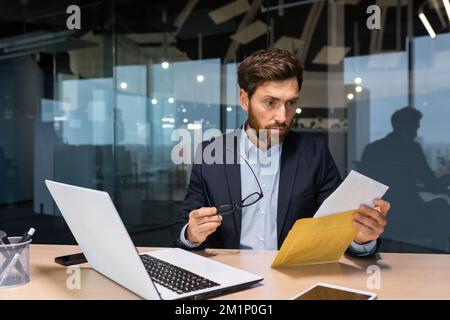 Ein schockierter junger Mann sitzt im Büro am Schreibtisch und hält einen Umschlag mit einem Brief und einer Brille in der Hand. Schlechte Nachrichten, Rechnung, Kreditvertrag. Stockfoto