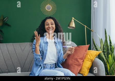 Porträt einer schönen Frau zu Hause, einer hispanischen Frau, die glücklich lächelt und mit dem Ergebnis des Schwangerschaftstests zufrieden in die Kamera schaut, auf dem Sofa im Wohnzimmer sitzt. Stockfoto