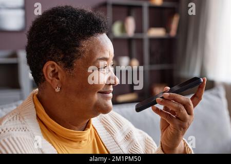 Seitliches Porträt einer lächelnden schwarzen Seniorin, die Sprachnachrichten per Smartphone zu Hause aufnimmt, Kopierbereich Stockfoto