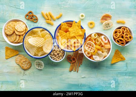 Salzige Snacks. Partyessen-Mix. Kartoffel- und Tortilla-Chips, Cracker und andere Vorspeisen in Schüsseln, über Kopf flach liegend auf blauem Hintergrund Stockfoto