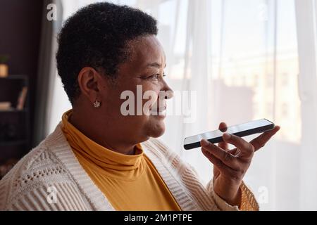 Seitliches Porträt einer schwarzen Seniorin, die Sprachnachrichten per Smartphone gegen das Fenster aufnimmt Stockfoto