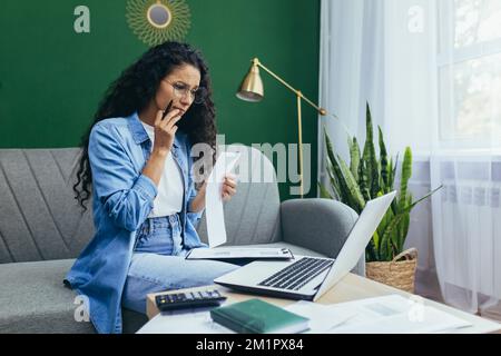 Seriöse und umsichtige Geschäftsfrau, die zu Hause mit Dokumenten arbeitet, hispanische Frau in legerer Kleidung, die bei der Arbeit ein Notebook benutzt und auf dem Sofa im Wohnzimmer mit Rechnungen und Verträgen sitzt. Stockfoto