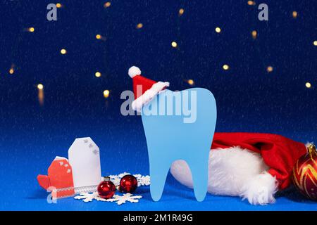 Weihnachtszahnheilkunde - Zahn mit weihnachtsmann-Hut, Fäustlinge auf blauem Hintergrund mit Schneefall. Stockfoto