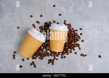 Zwei Einweg-Pappbecher für heißen Kaffee liegen auf Kaffeebohnen auf hellem Betonhintergrund Stockfoto