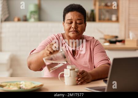 In warmen Farbtönen gehaltenes Porträt einer schwarzen Seniorin, die während des Frühstücks Tee in einer gemütlichen Küche macht Stockfoto