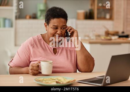 Warmes Porträt einer schwarzen Seniorin, die am Telefon spricht, während sie morgens frühstückt Stockfoto