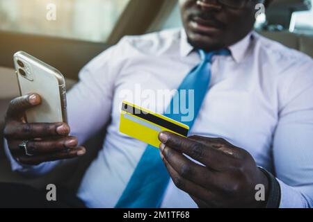 Der afroamerikanische Geschäftsmann kauft online über das Telefon, während er auf dem Rücksitz des Autos sitzt und eine Kreditkarte verwendet Stockfoto