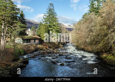 Die Wasserfälle von Dochart blicken flussabwärts von der Brücke und zeigen weißes Wasser, Felsen, einen Bungalow am Flussufer, Kiefern und schneebedeckte Berge. Stockfoto